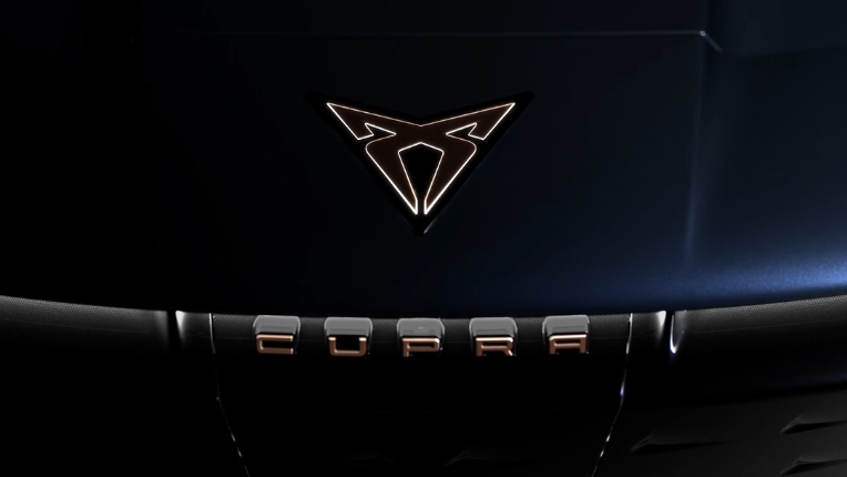 Detailný pohľad na kapotu auta, kde je umiestnené logo značky CUPRA. Logo má charakteristický tvar v tmavej kovovej farbe. Na maske je tiež vidieť nápis "CUPRA".