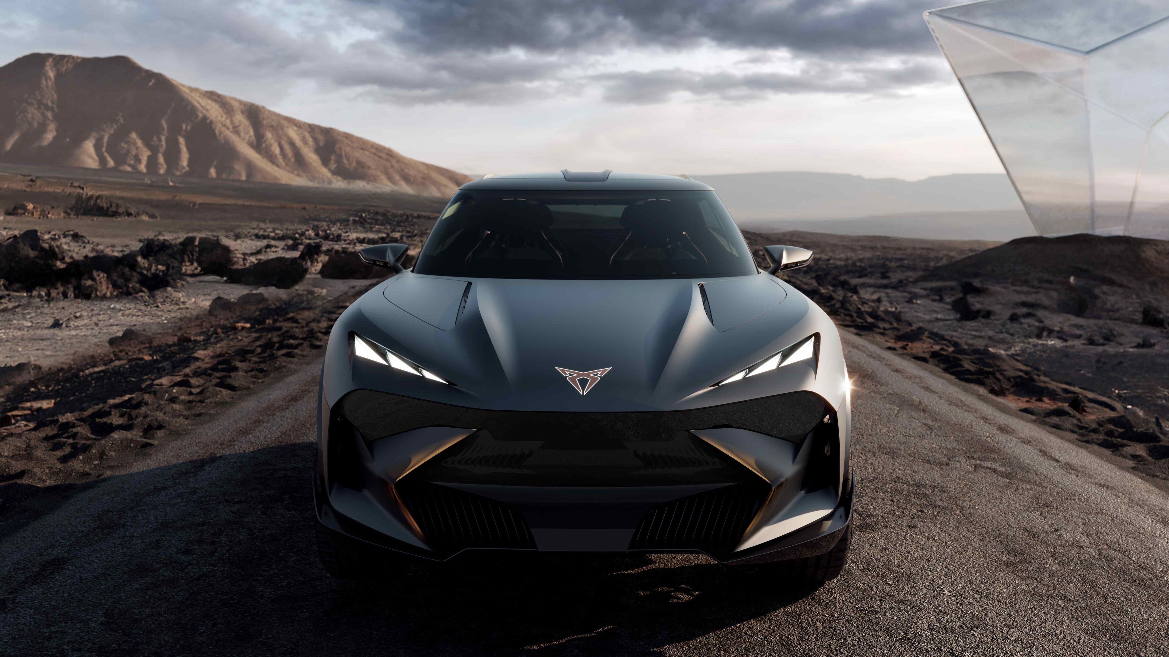 Futuristické športové auto s moderným dizajnom stojí na ceste v púštnej krajine. Nad autom je zamračená obloha a v pozadí sú skalnaté hory. Auto má agresívne línie, tmavý lak, subtílne svetlomety a výraznú prednú masku.