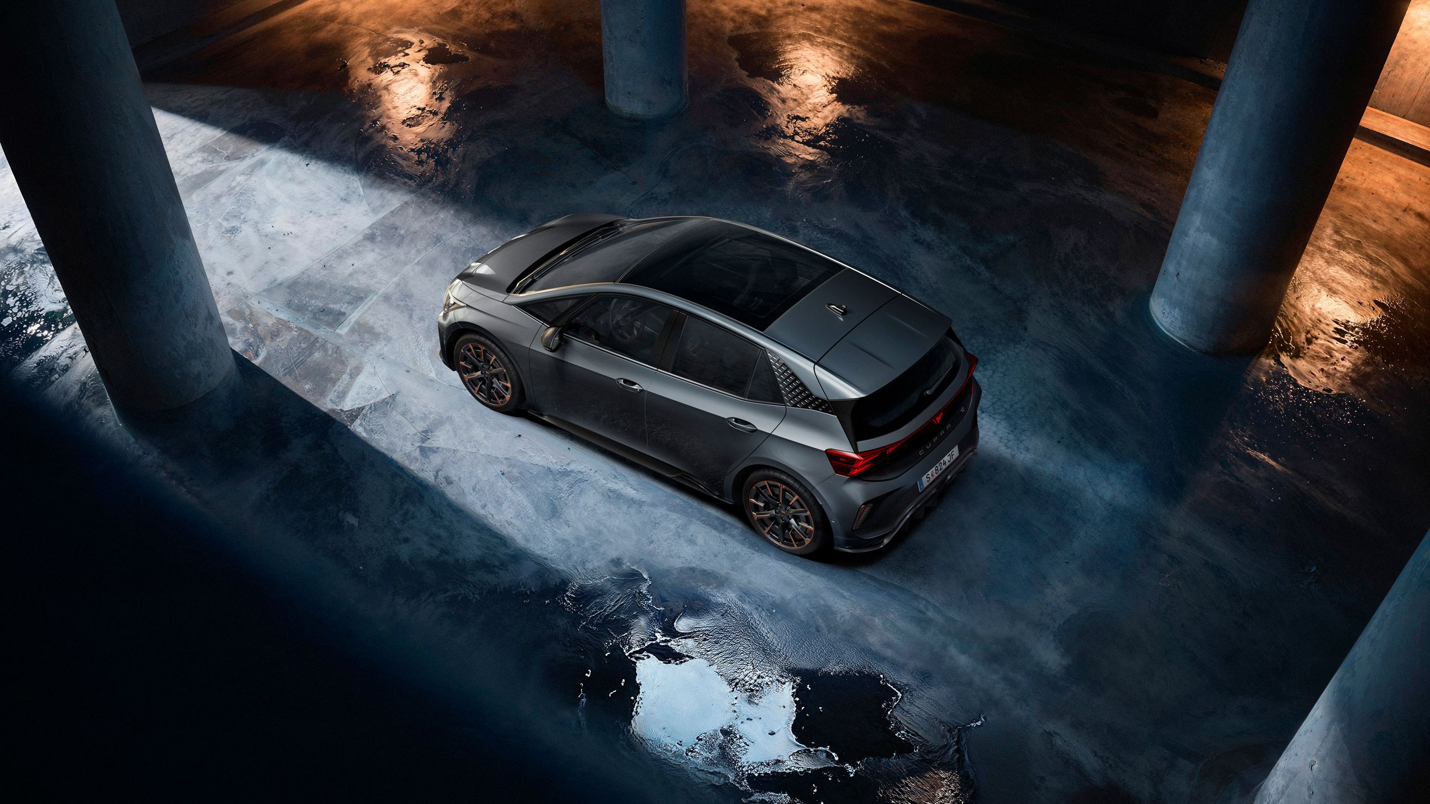 Sivý CUPRA Born stojí v tmavej podzemnej garáži. Fotografia je nasnímaná z vtáčej perspektívy a zachytáva odlesky svetla na mokrej betónovej podlahe. Scénu osvetľuje nepriame svetlo, ktoré vytvára dramatickú atmosféru.