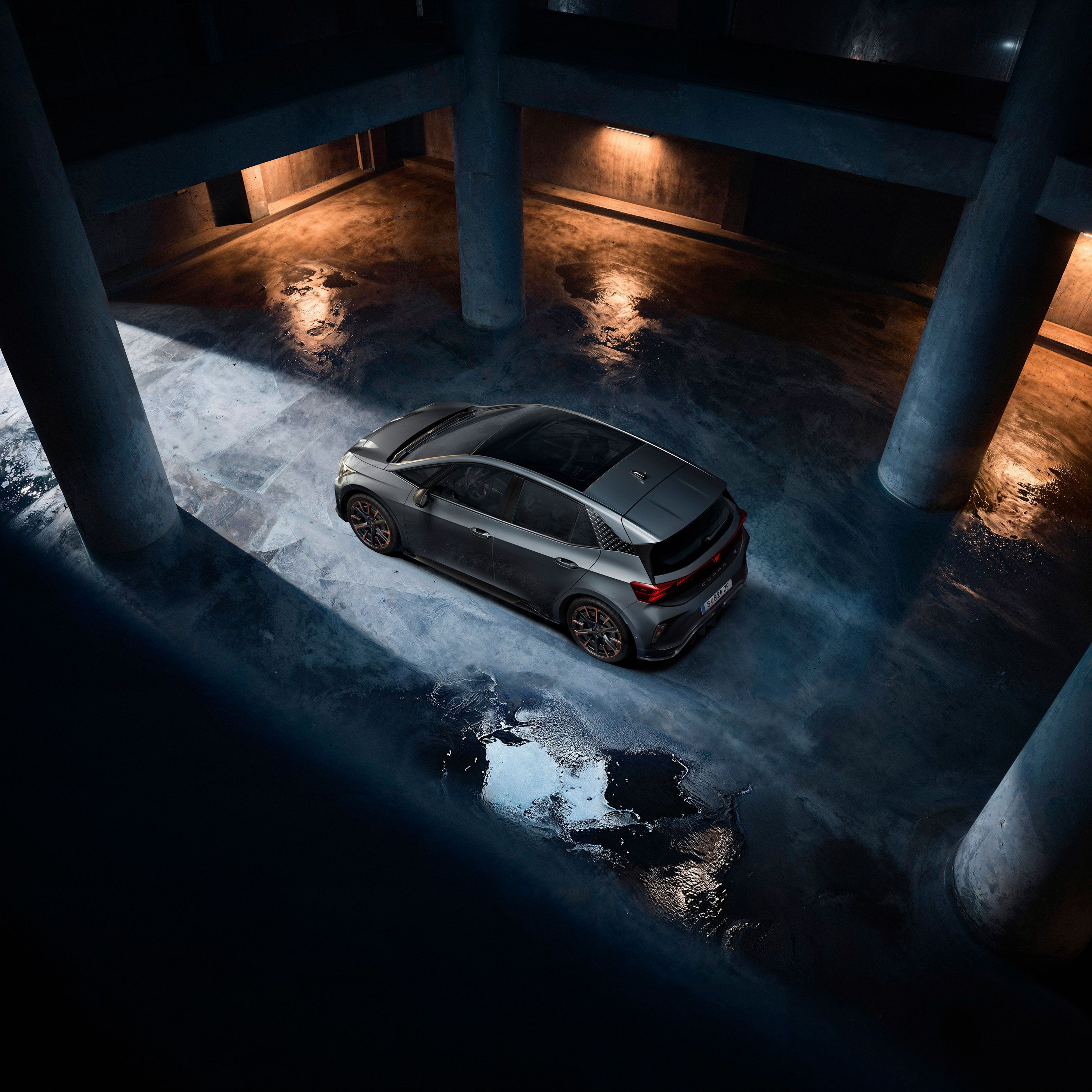Sivý CUPRA Born stojí v tmavej podzemnej garáži. Fotografia je nasnímaná z vtáčej perspektívy a zachytáva odlesky svetla na mokrej betónovej podlahe. Scénu osvetľuje nepriame svetlo, ktoré vytvára dramatickú atmosféru.