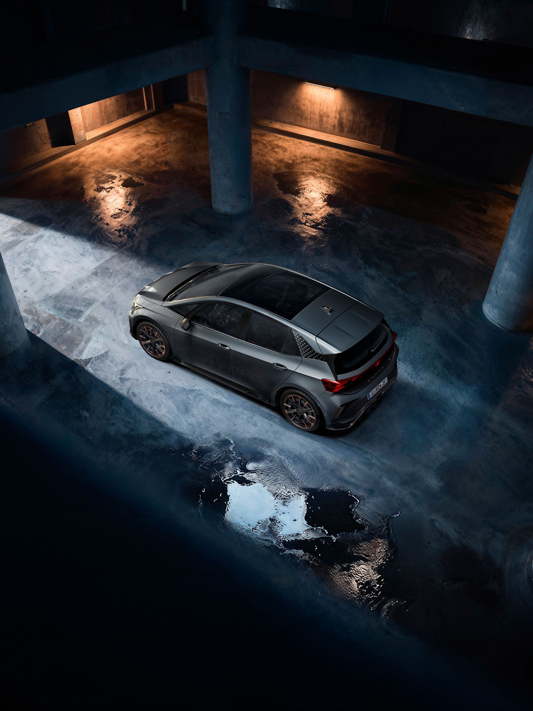 Sivý CUPRA Born stojí v tmavej podzemnej garáži. Fotografia je nasnímaná z vtáčej perspektívy a zachytáva odlesky svetla na mokrej betónovej podlahe. Scénu osvetľuje nepriame svetlo, ktoré vytvára dramatickú atmosféru.