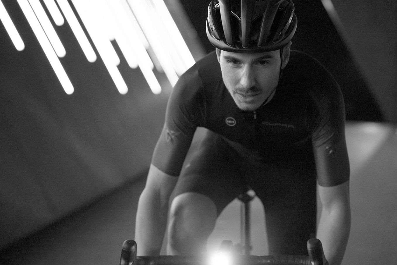 Cyklista v športovom oblečení je zachytený pri jazde na bicykli v prostredí s výraznými svetelnými prvkami na stene.