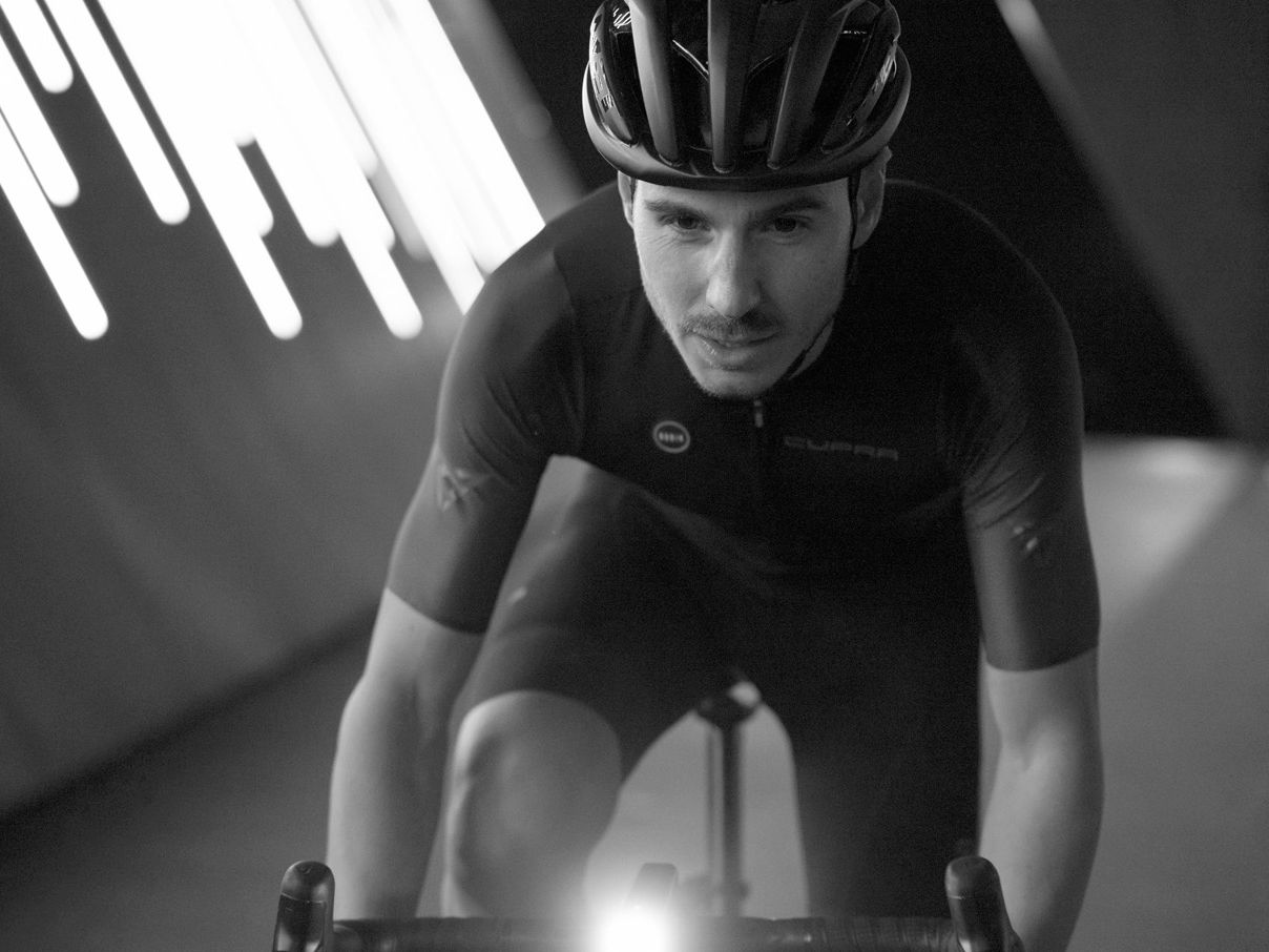 Cyklista v športovom oblečení je zachytený pri jazde na bicykli v prostredí s výraznými svetelnými prvkami na stene.