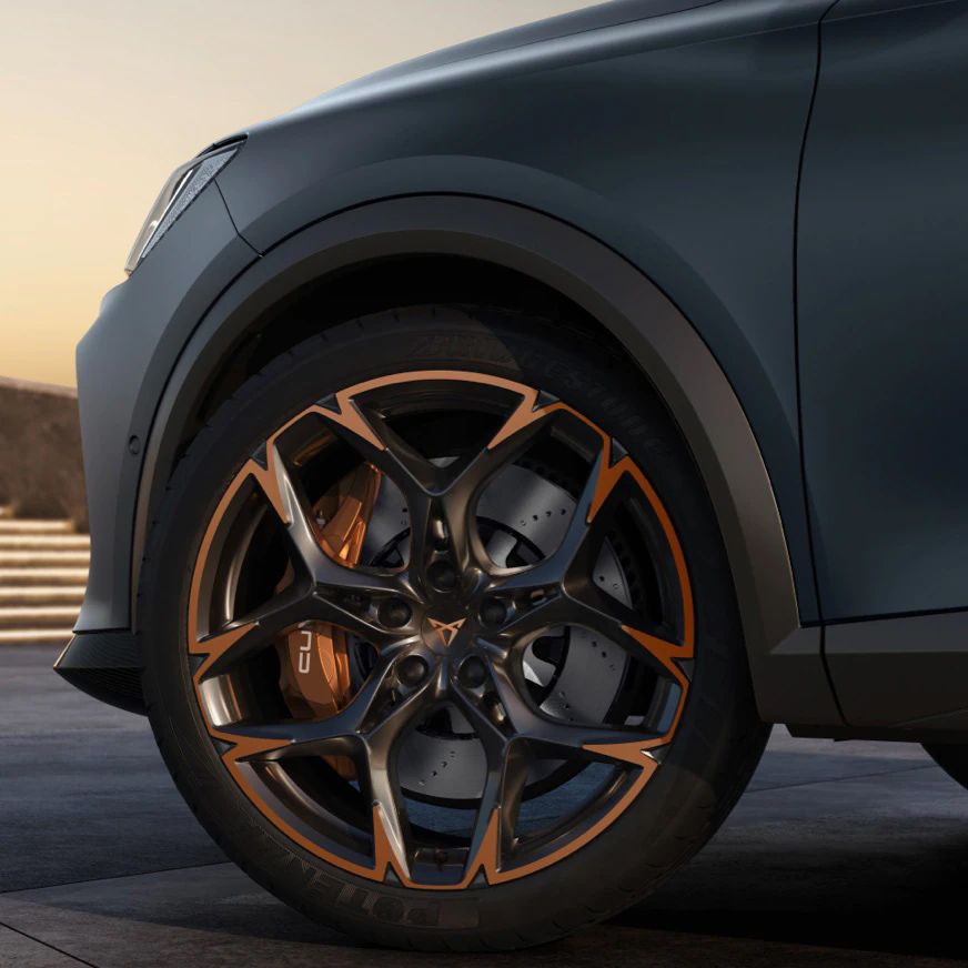 Detailný záber na športové koleso automobilu s čiernym diskom s bronzovými prvkami. Pozadie je rozmazané a zahŕňa schody a nejasné objekty. Koleso je súčasťou tmavošedého auta.