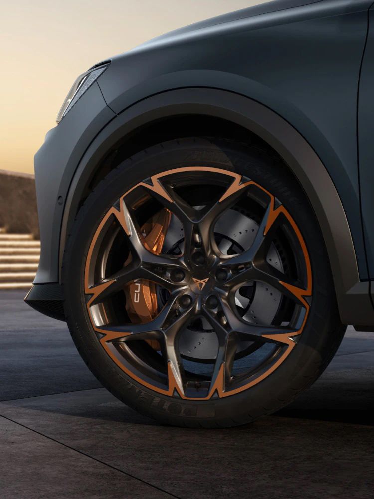 Detailný záber na športové koleso automobilu s čiernym diskom s bronzovými prvkami. Pozadie je rozmazané a zahŕňa schody a nejasné objekty. Koleso je súčasťou tmavošedého auta.