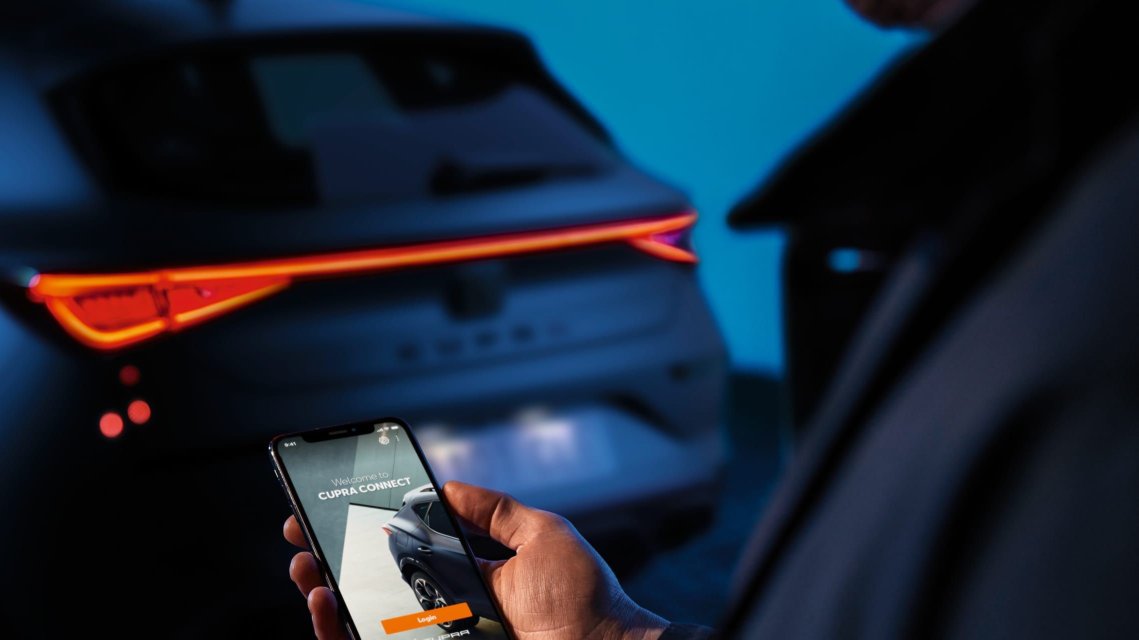Muž drží mobilný telefón, na ktorom je zobrazená aplikácia Cupra Connect. V pozadí sa nachádza auto značky Cupra s rozsvietenými zadnými svetlami.