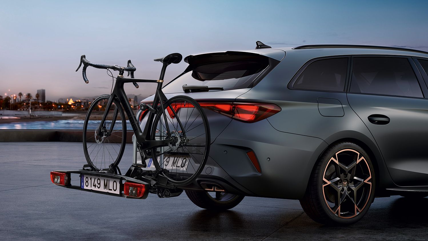 Automobil je zaparkovaný na pobreží počas západu slnka. Na zadnej časti má nainštalovaný nosič bicyklov na ťažné zariadenie. V pozadí je vidieť mestské budovy, palmy a pobrežie.