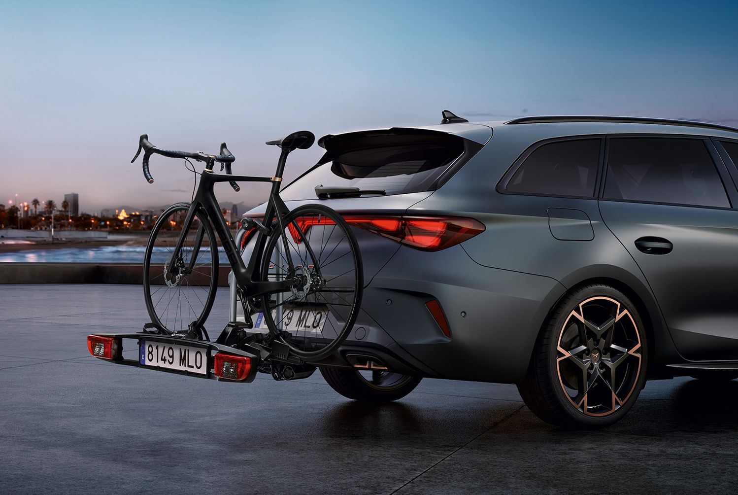 Automobil je zaparkovaný na pobreží počas západu slnka. Na zadnej časti má nainštalovaný nosič bicyklov na ťažné zariadenie. V pozadí je vidieť mestské budovy, palmy a pobrežie.