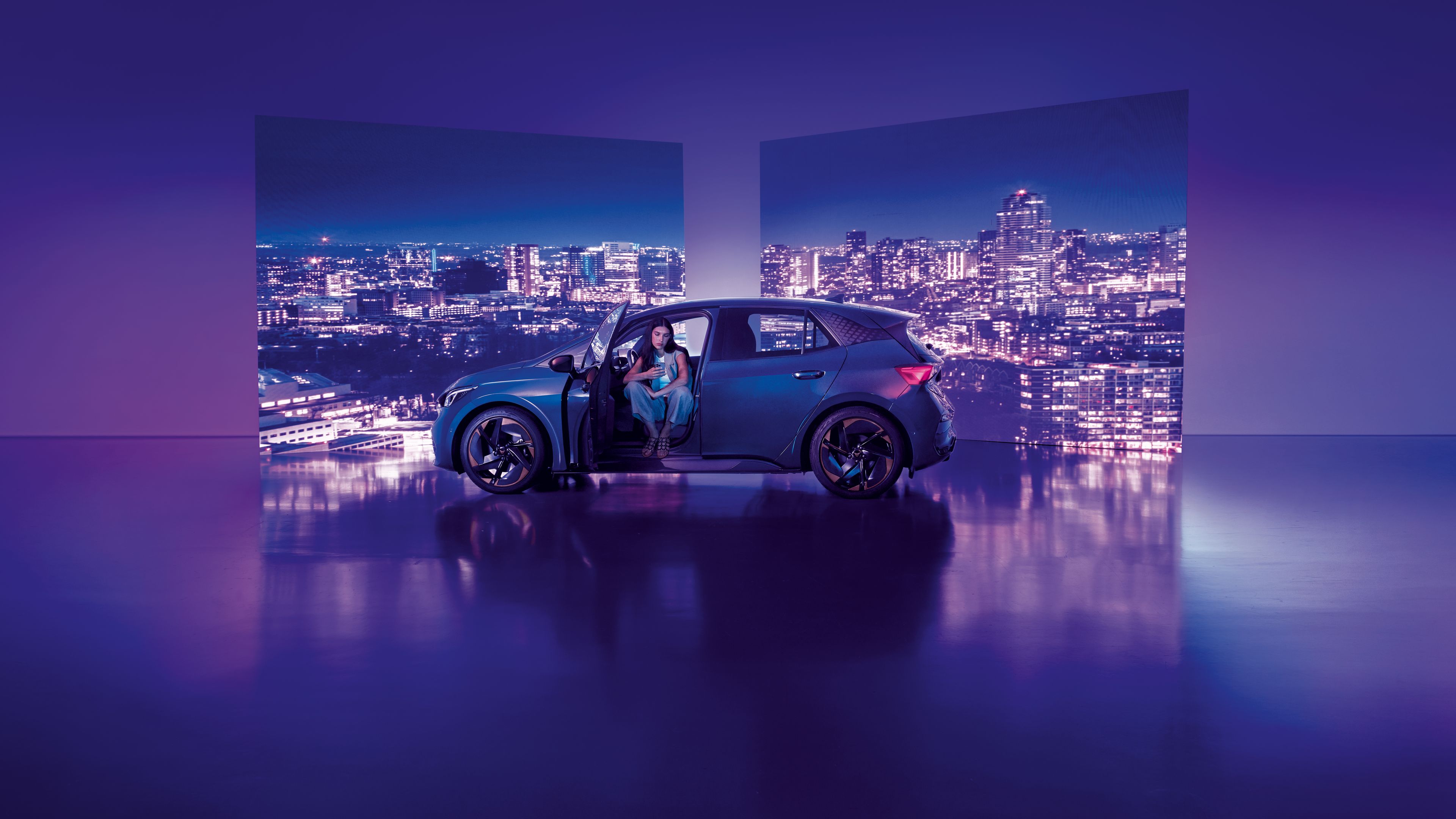 Pozadie tvoria dva veľké billboardy s panoramatickým výhľadom na nočné mesto, pred ktorými stojí moderné auto s otvorenými dverami. Žena sedí v aute na mieste vodiča, interiér je osvetlený jemným svetlom.