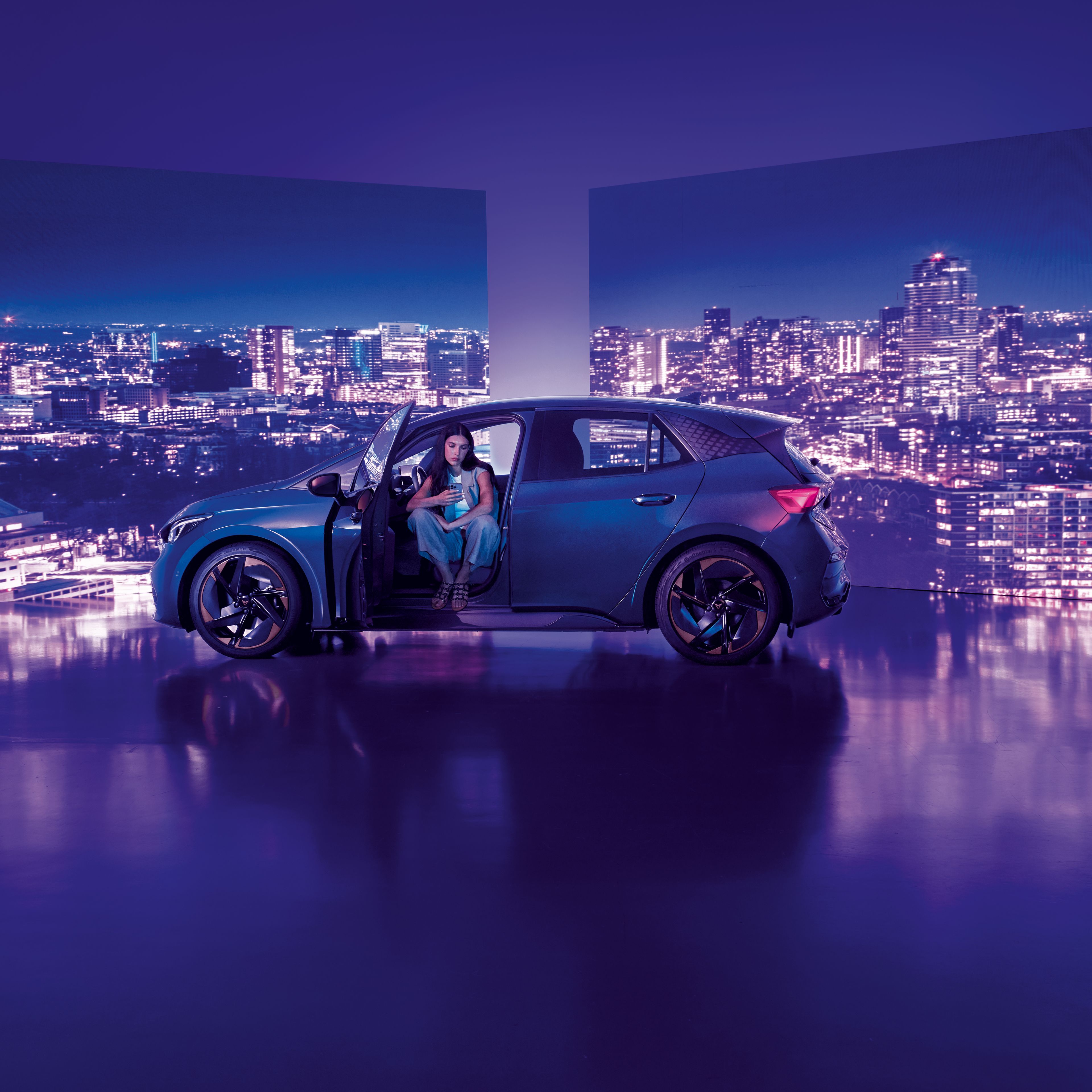 Pozadie tvoria dva veľké billboardy s panoramatickým výhľadom na nočné mesto, pred ktorými stojí moderné auto s otvorenými dverami. Žena sedí v aute na mieste vodiča, interiér je osvetlený jemným svetlom.