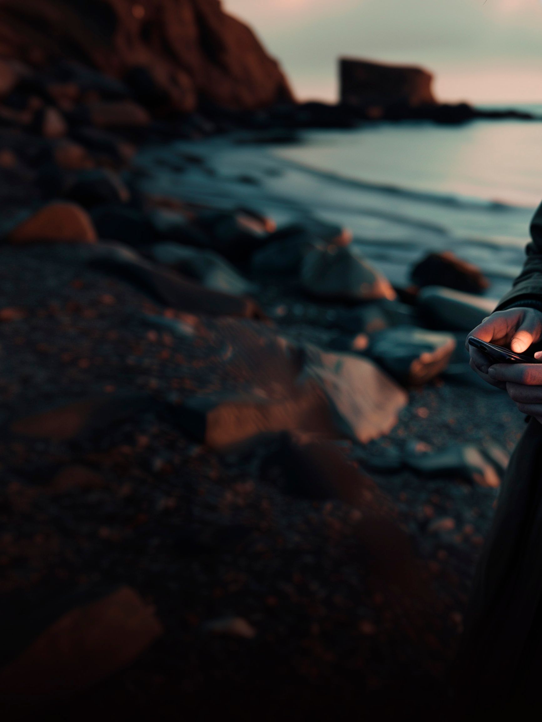 Osoba stojí na kamenistom brehu mora, drží telefón a pozerá sa na displej. V pozadí je skalnaté pobrežie počas súmraku.