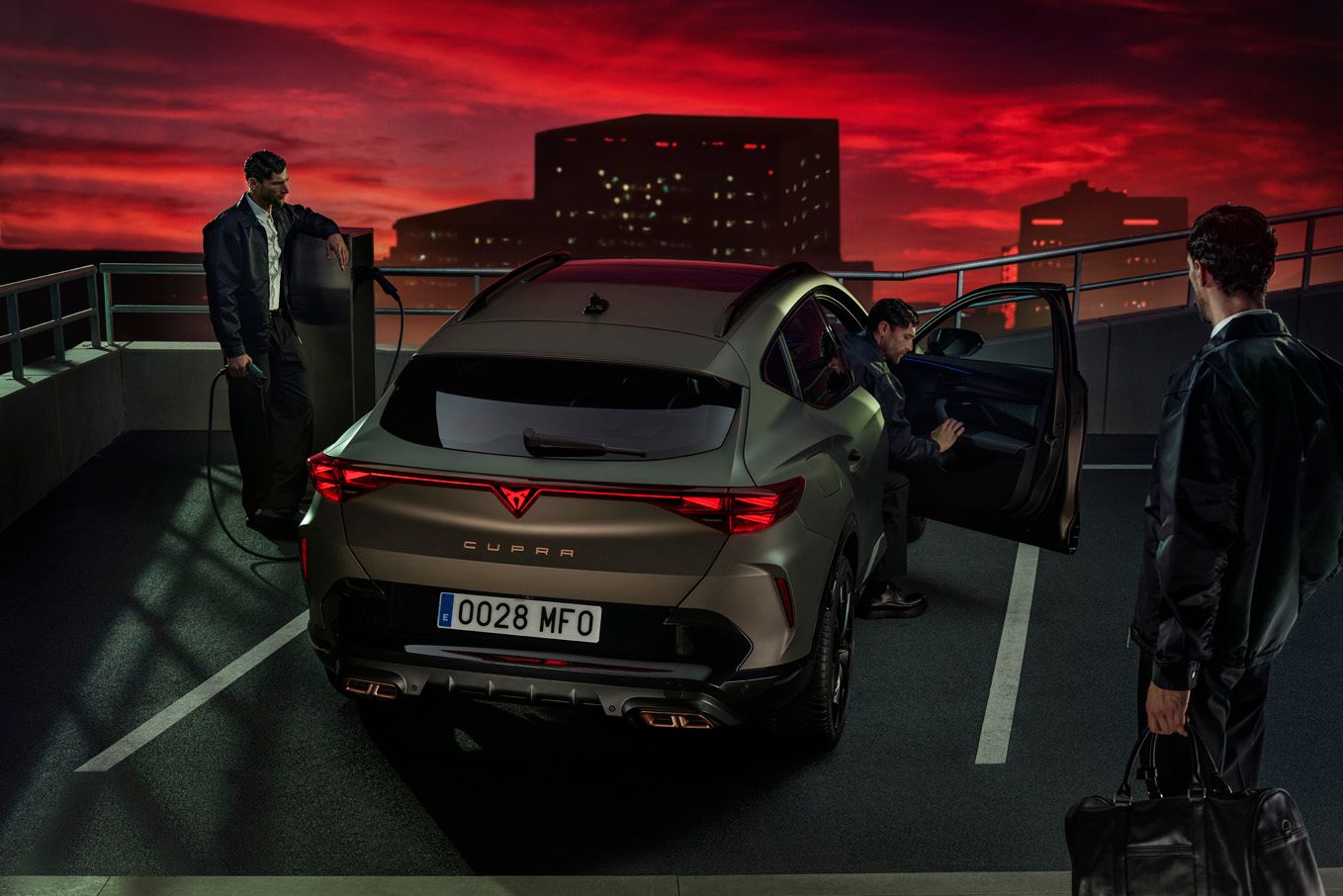 Dvaja muži stoja pri aute značky CUPRA a jeden vystupuje z vozidla. Jeden z mužov drží nabíjací konektor, iný tašku zbalenú na víkend. Na oblohe je západ slnka, v pozadí sú osvetlené budovy.