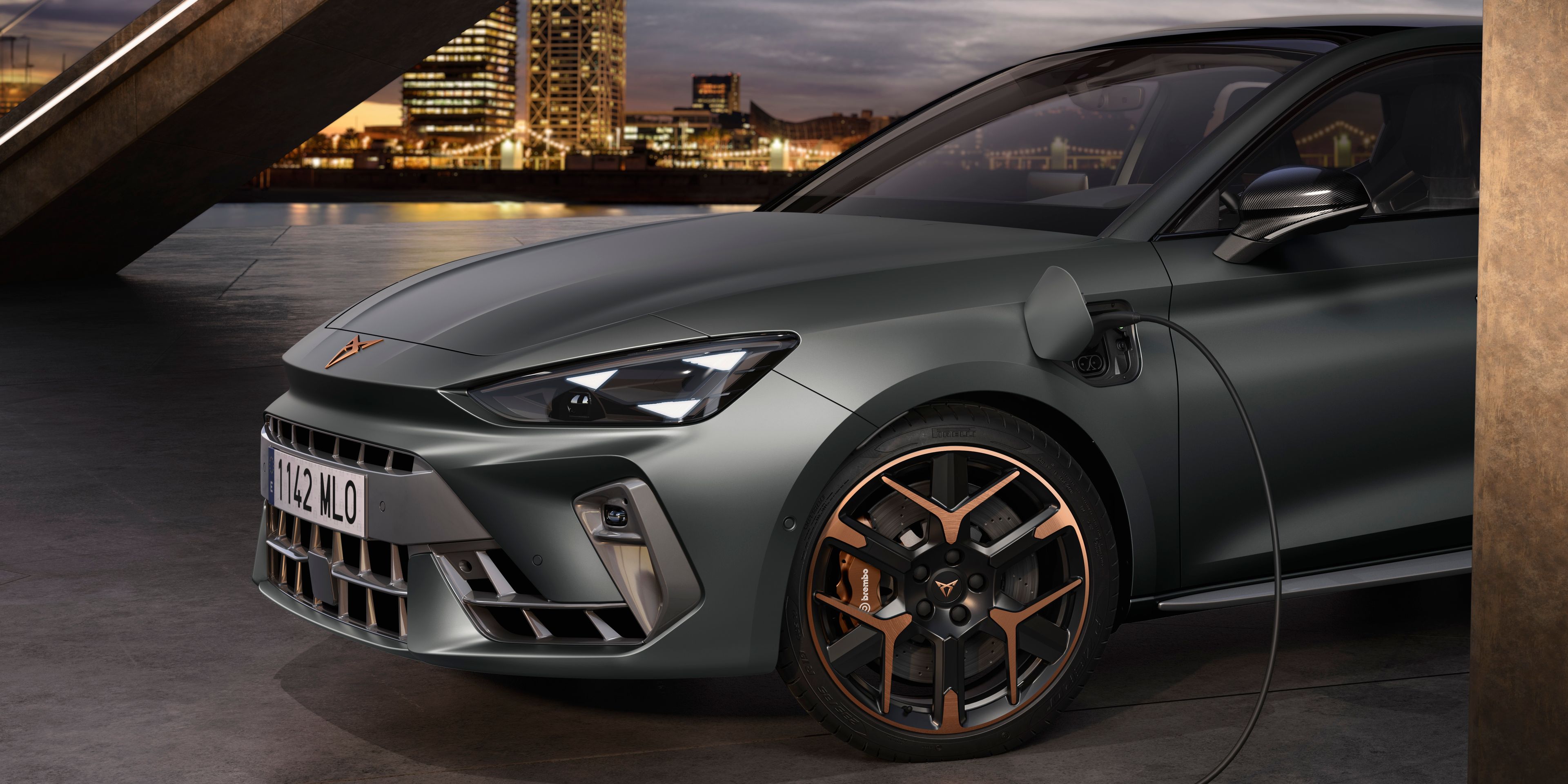 Elektrický automobil Cupra Leon je pripojený k nabíjaciemu káblu v modernom mestskom prostredí. V pozadí je vidieť betónovú lávku pre peších a mrakodrapy. Auto je šedej farby s bronzovými detailmi na kolesách.