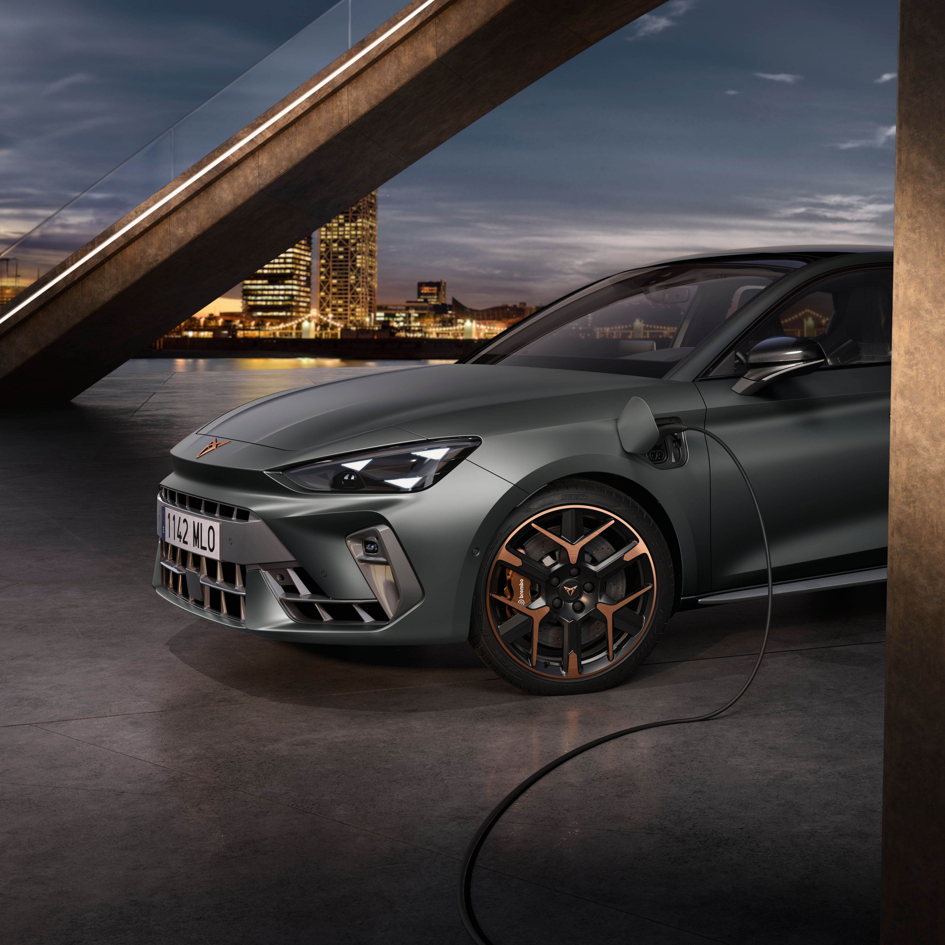 Elektrický automobil Cupra Leon je pripojený k nabíjaciemu káblu v modernom mestskom prostredí. V pozadí je vidieť betónovú lávku pre peších a mrakodrapy. Auto je šedej farby s bronzovými detailmi na kolesách.