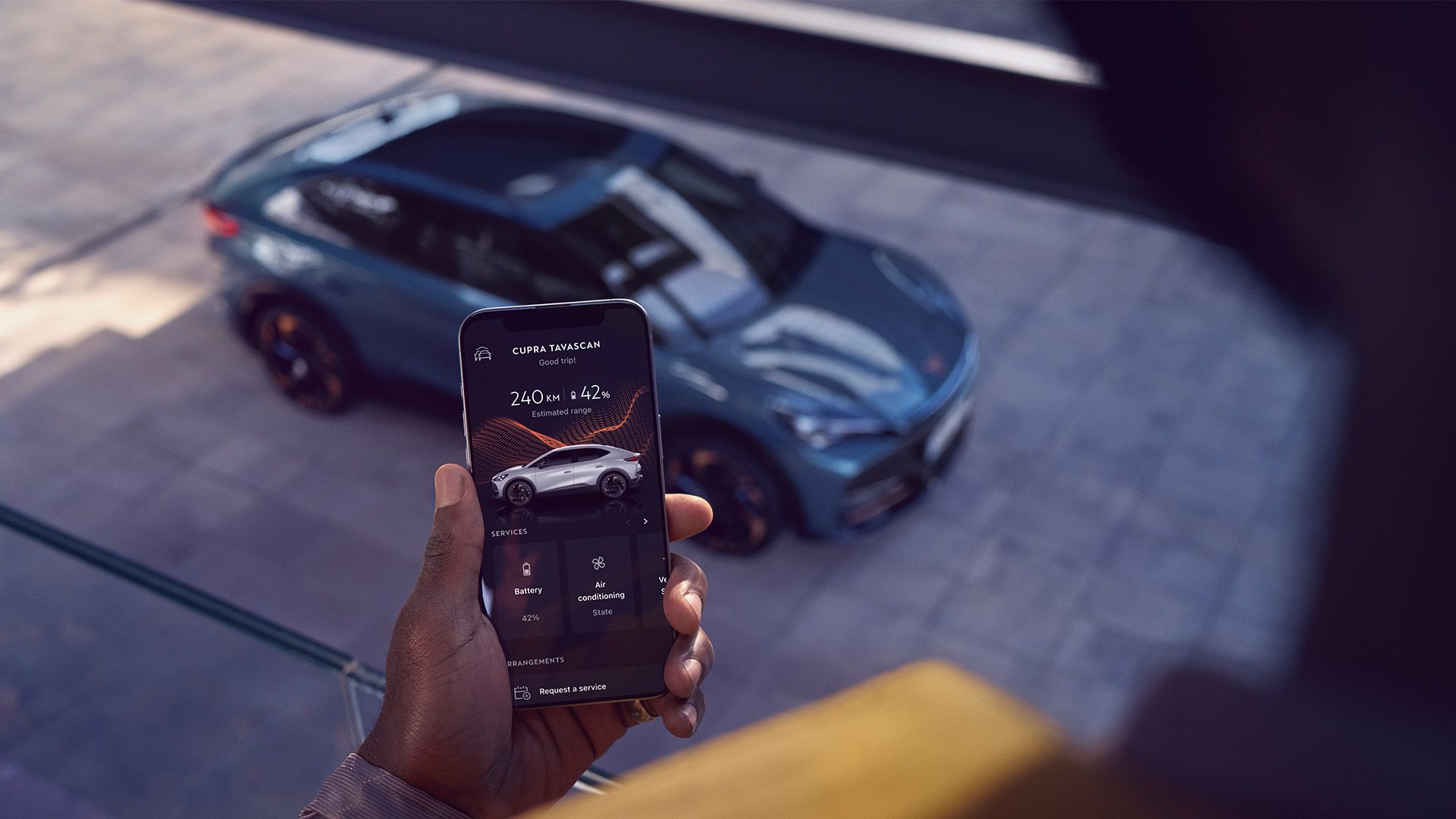 Muž drží mobilný telefón a pomocou aplikácie ovláda auto zaparkované von. Aplikácia ukazuje informácie o stave vozidla. Auto je moderné a tmavo sivé.