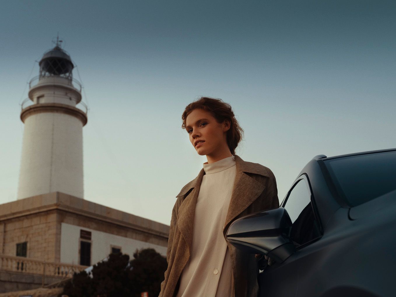 Žena stojí vedľa auta pred majákom za súmraku. Má na sebe kabát a svetlý rolák. Pozadie tvorí obloha jemne zafarbená do modra.