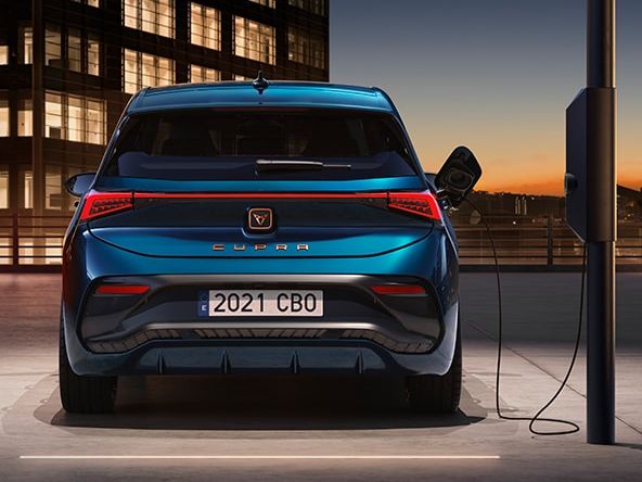 Auto značky CUPRA je zaparkované pri nabíjacej stanici a je pripojené. Je to súčasť moderného mestského prostredia, ktoré zdôrazňuje technologický pokrok a udržateľnosť. Na pozadí je budova pri západe slnka.