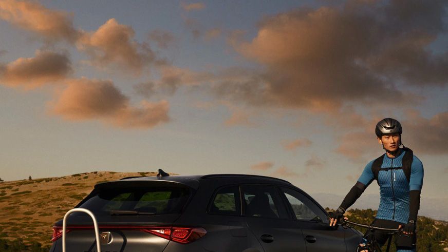 Muž oblečený v cyklistickom oblečení stojí pri zaparkovanom aute na horskej ceste počas západu slnka.