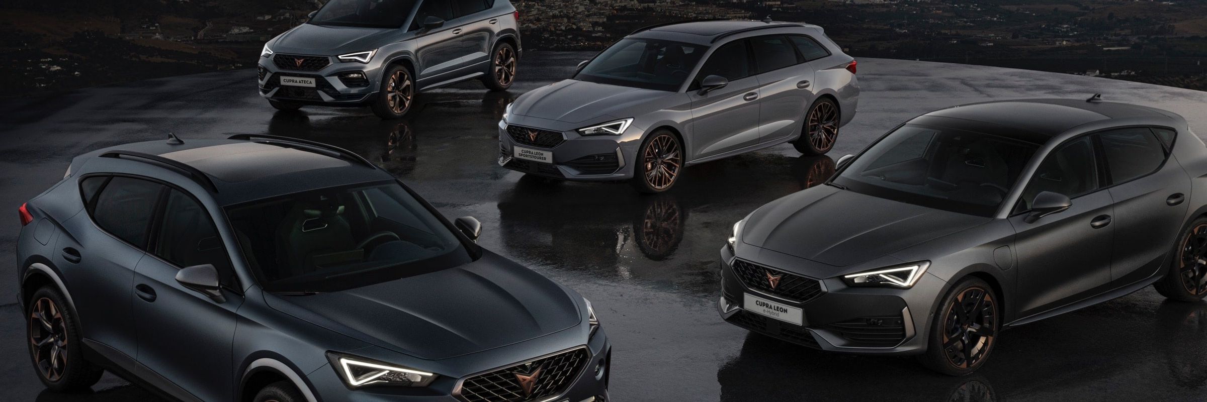 Štyri moderné tmavosivé športové automobily CUPRA zaparkované na mokrej vozovke pri západe slnka. V pozadí je vidieť osvetlené mesto. Všetky autá majú športový dizajn.
