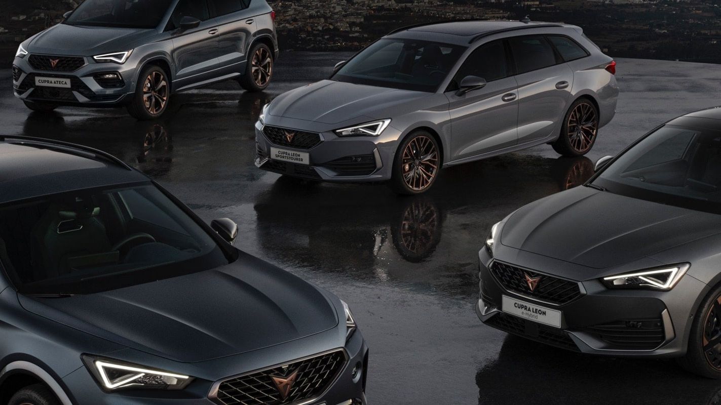 Štyri moderné tmavosivé športové automobily CUPRA zaparkované na mokrej vozovke pri západe slnka. V pozadí je vidieť osvetlené mesto. Všetky autá majú športový dizajn.