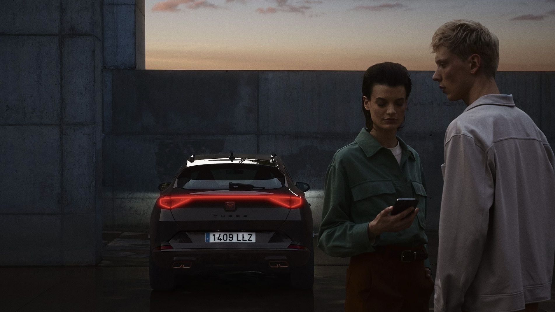 Na obrázku sú dvaja ľudia stojaci za zaparkovaným vozidlom počas súmraku. Jeden z nich drží mobilný telefón. Auto je športového typu a má zapnuté zadné svetlá.