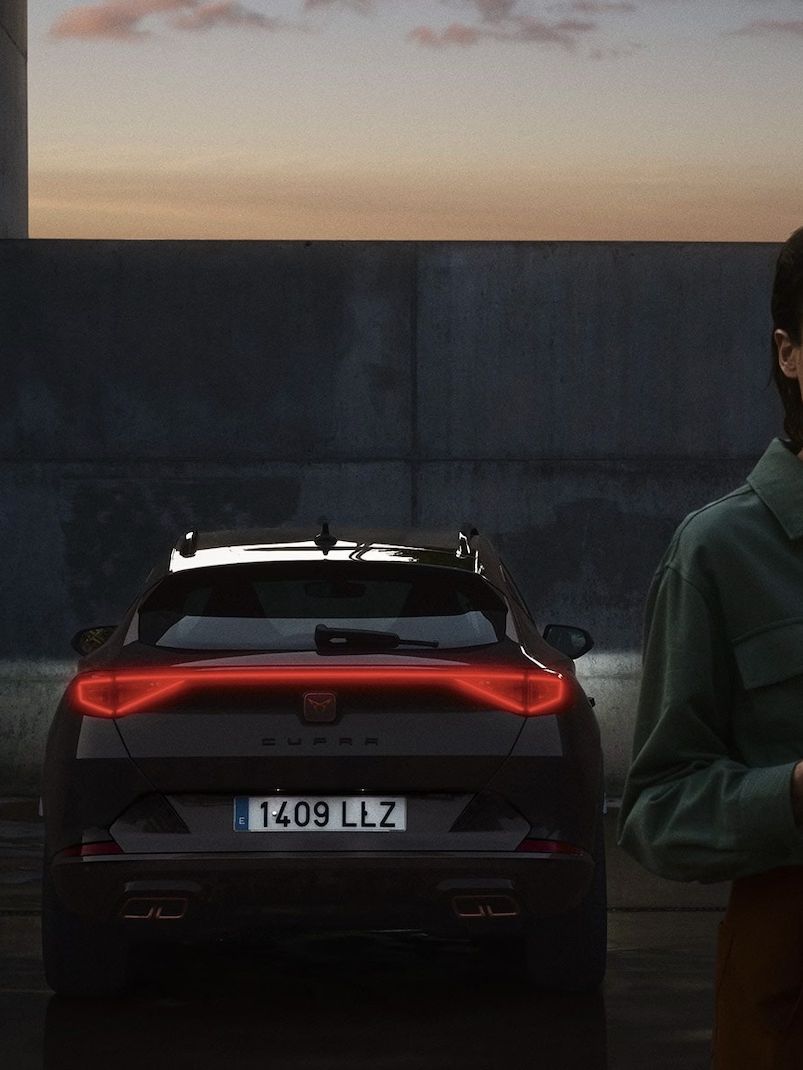 Na obrázku sú dvaja ľudia stojaci za zaparkovaným vozidlom počas súmraku. Jeden z nich drží mobilný telefón. Auto je športového typu a má zapnuté zadné svetlá.