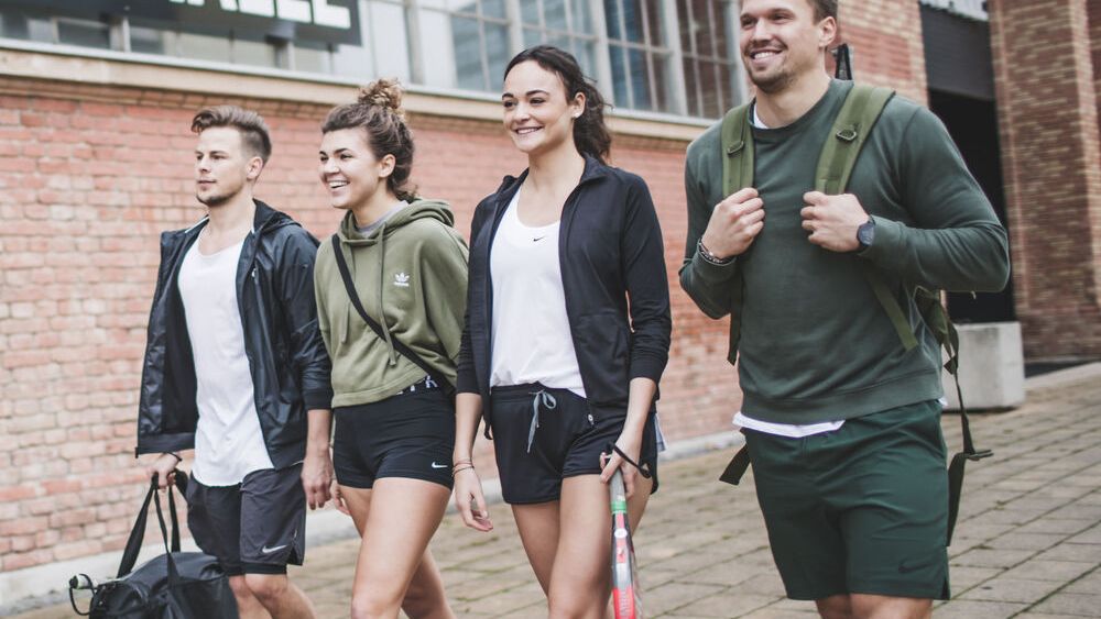 Štyria ľudia oblečení v športovom oblečení a so športovými taškami kráčajú po chodníku vedľa tehlovej budovy s nápisom "BOX HALLE". Dvaja z nich majú v rukách padelové rakety.