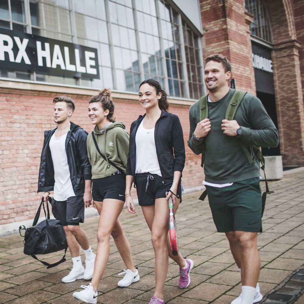 Štyria ľudia oblečení v športovom oblečení a so športovými taškami kráčajú po chodníku vedľa tehlovej budovy s nápisom "BOX HALLE". Dvaja z nich majú v rukách padelové rakety.