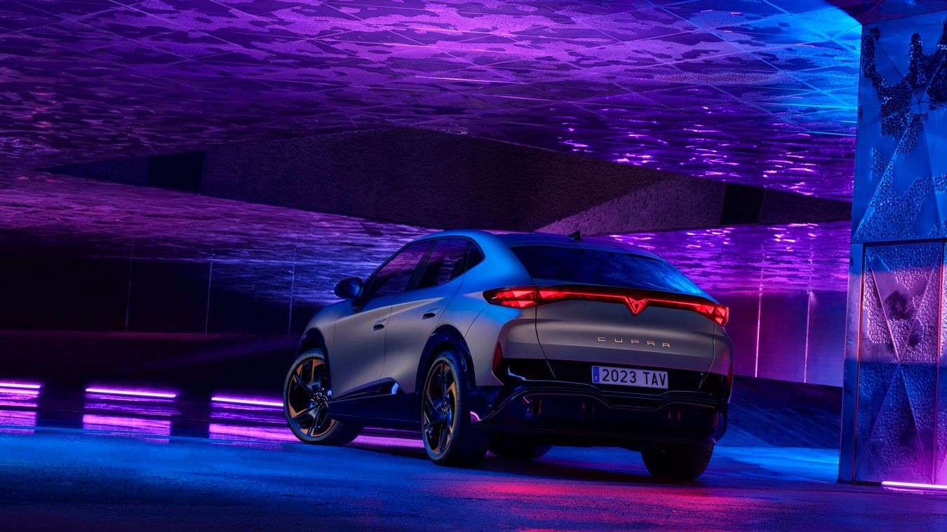 Elegantné športové auto CUPRA zaparkované v modernom, futuristickom osvetlenom prostredí. Auto má tmavosivú farbu s výraznými červenými zadnými svetlami. Štýlový dizajn zvýrazňujú čierne kolesá s bronzovými prvkami.