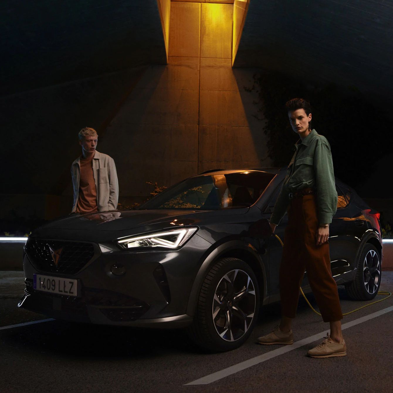 Dve osoby stoja vedľa tmavého moderného SUV, ktorý je zaparkovaný pod osvetleným nadjazdom.