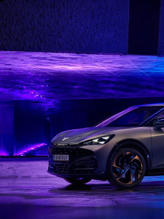 Tmavosivé moderné CUPRA Tavascan je zaparkované v osvetlenom podzemnom priestore s fialovým a modrým osvetlením. Jeho dizajn je športový a aerodynamický. Pozadie pôsobí futuristicky.