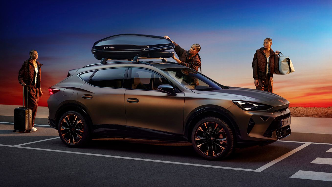 Moderné SUV je zaparkované vedľa cesty pri západe slnka, ľudia nakladajú batožinu na strechu a do kufra auta. Športovo vyzerajúce auto má tmavé metalické lakovanie a elegantný dizajn.