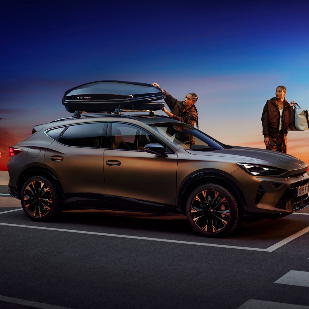 Moderné SUV je zaparkované vedľa cesty pri západe slnka, ľudia nakladajú batožinu na strechu a do kufra auta. Športovo vyzerajúce auto má tmavé metalické lakovanie a elegantný dizajn.