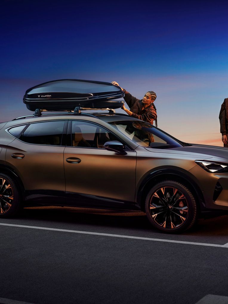 Moderné SUV je zaparkované vedľa cesty pri západe slnka, ľudia nakladajú batožinu na strechu a do kufra auta. Športovo vyzerajúce auto má tmavé metalické lakovanie a elegantný dizajn.