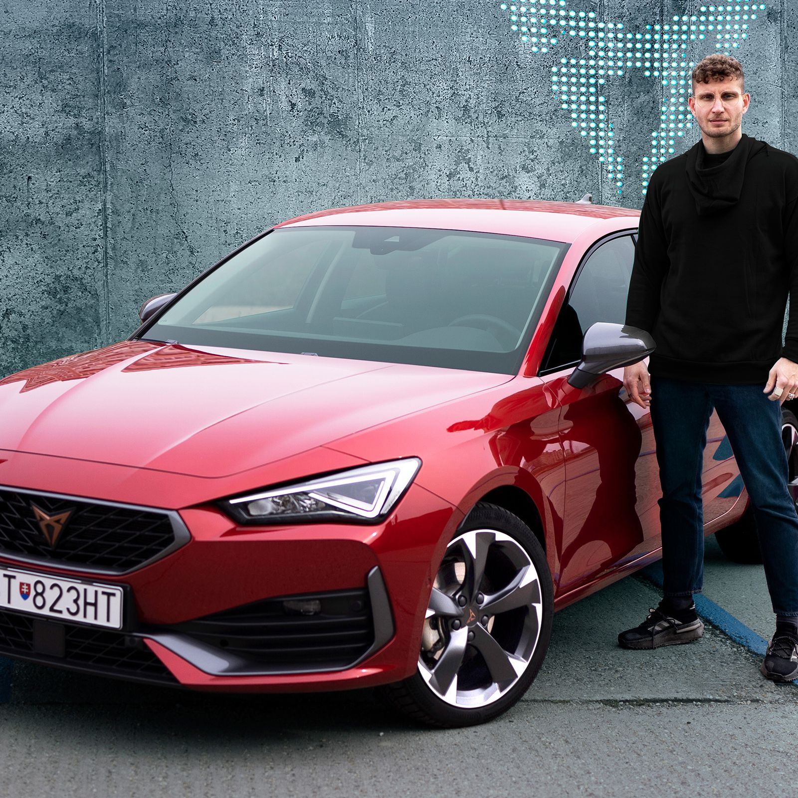 Muž stojí vedľa červeného športového auta. Auto je zaparkované na betónovom povrchu. Muž má na sebe tmavé oblečenie. Na betónovej stene za vozidlom je logo CUPRA.