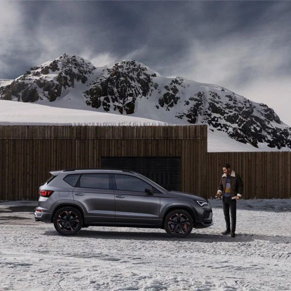 Šedé SUV CUPRA stojí pri drevenej budove v zasneženej horskej krajine. Muž stojí vedľa auta. V pozadí sú zasnežené hory.