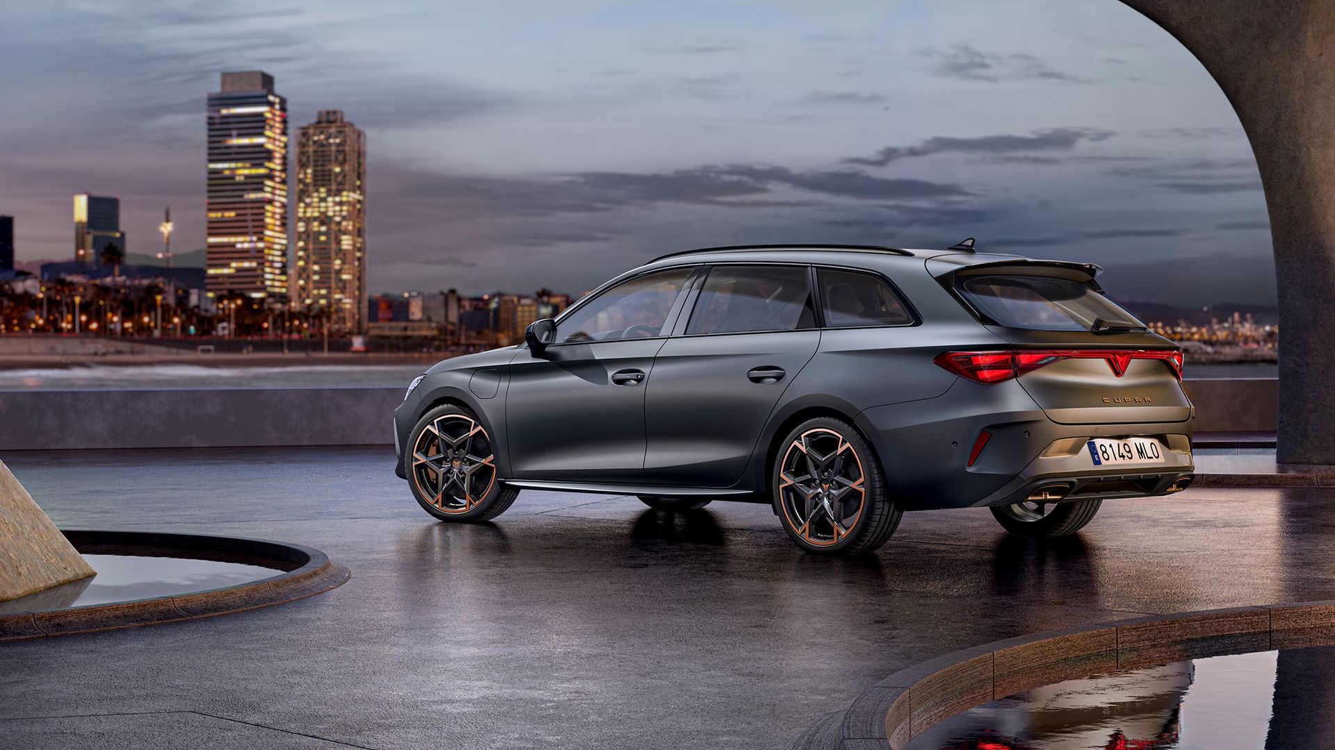 Moderné sivé CUPRA Leon Sportstourer zaparkovaný na voľnej ploche pri mori. V pozadí je výhľad na panorámu mesta s vysokými budovami. Automobil má sofistikovaný dizajn a športové prvky. Scéna je zachytená za súmraku.