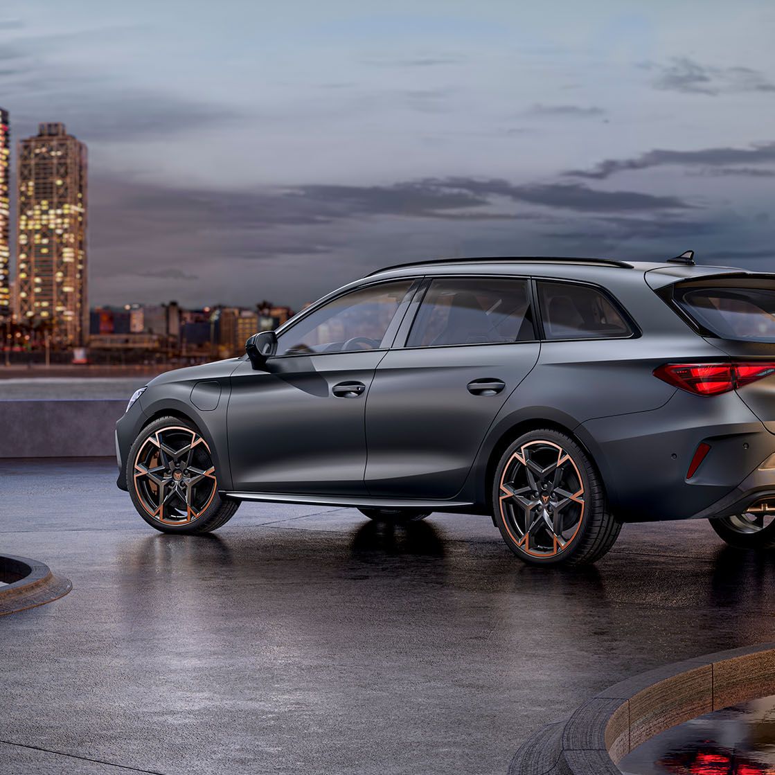 Moderné sivé CUPRA Leon Sportstourer zaparkovaný na voľnej ploche pri mori. V pozadí je výhľad na panorámu mesta s vysokými budovami. Automobil má sofistikovaný dizajn a športové prvky. Scéna je zachytená za súmraku.