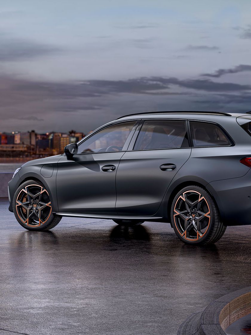 Moderné sivé CUPRA Leon Sportstourer zaparkovaný na voľnej ploche pri mori. V pozadí je výhľad na panorámu mesta s vysokými budovami. Automobil má sofistikovaný dizajn a športové prvky. Scéna je zachytená za súmraku.