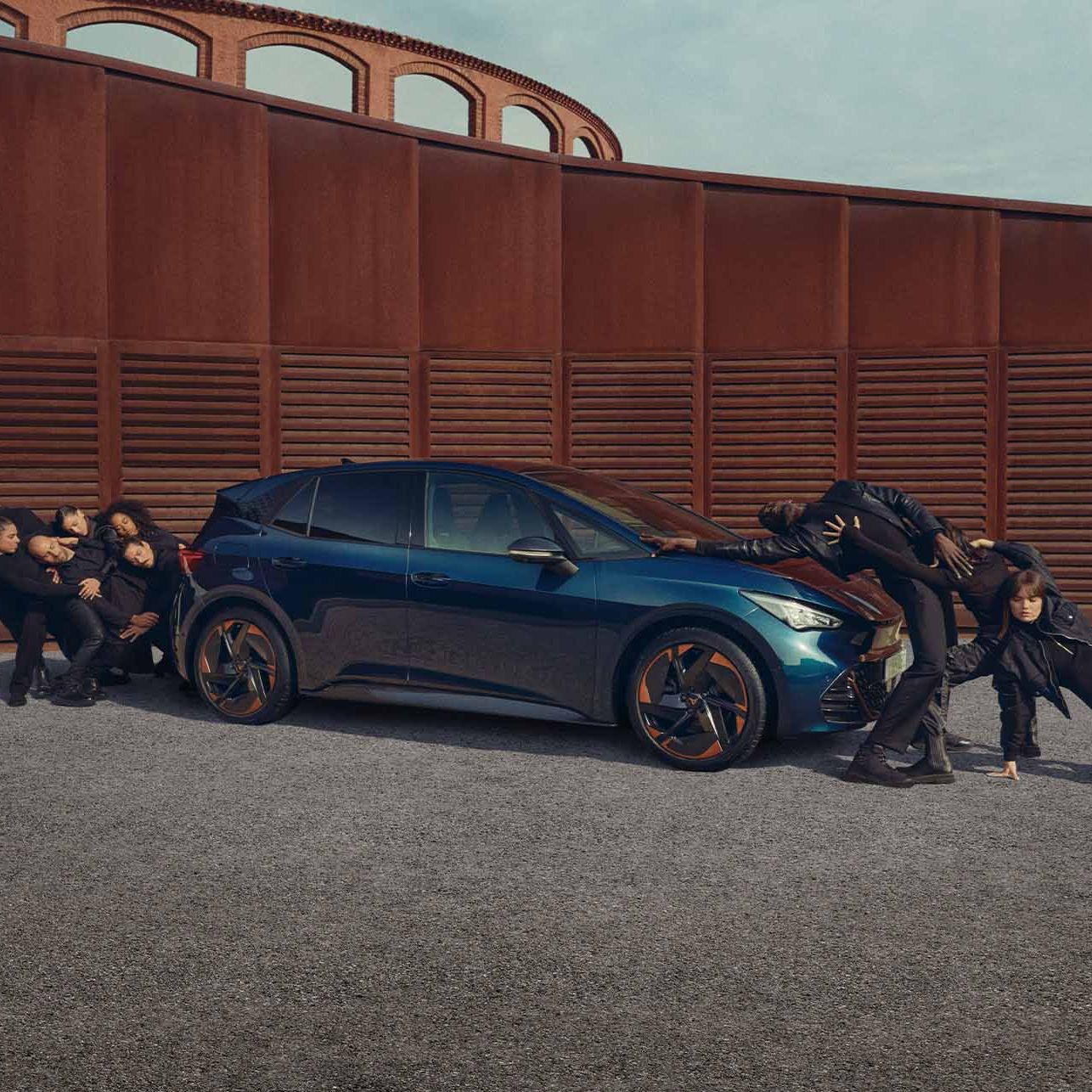 Skupina ľudí v čiernom umelecky stvárňuje pohyb pri vozidle pred kovovou cor-tenovou stenou. Niektorí členovia skupiny držia auto z boku, zatiaľ čo ostatní ho tlačia zo zadnej časti. Scéna sa odohráva na šedivej štrkovej ploche.