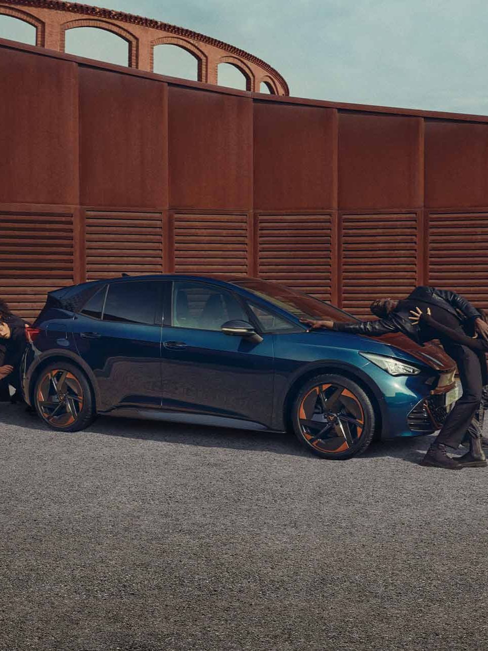 Skupina ľudí v čiernom umelecky stvárňuje pohyb pri vozidle pred kovovou cor-tenovou stenou. Niektorí členovia skupiny držia auto z boku, zatiaľ čo ostatní ho tlačia zo zadnej časti. Scéna sa odohráva na šedivej štrkovej ploche.