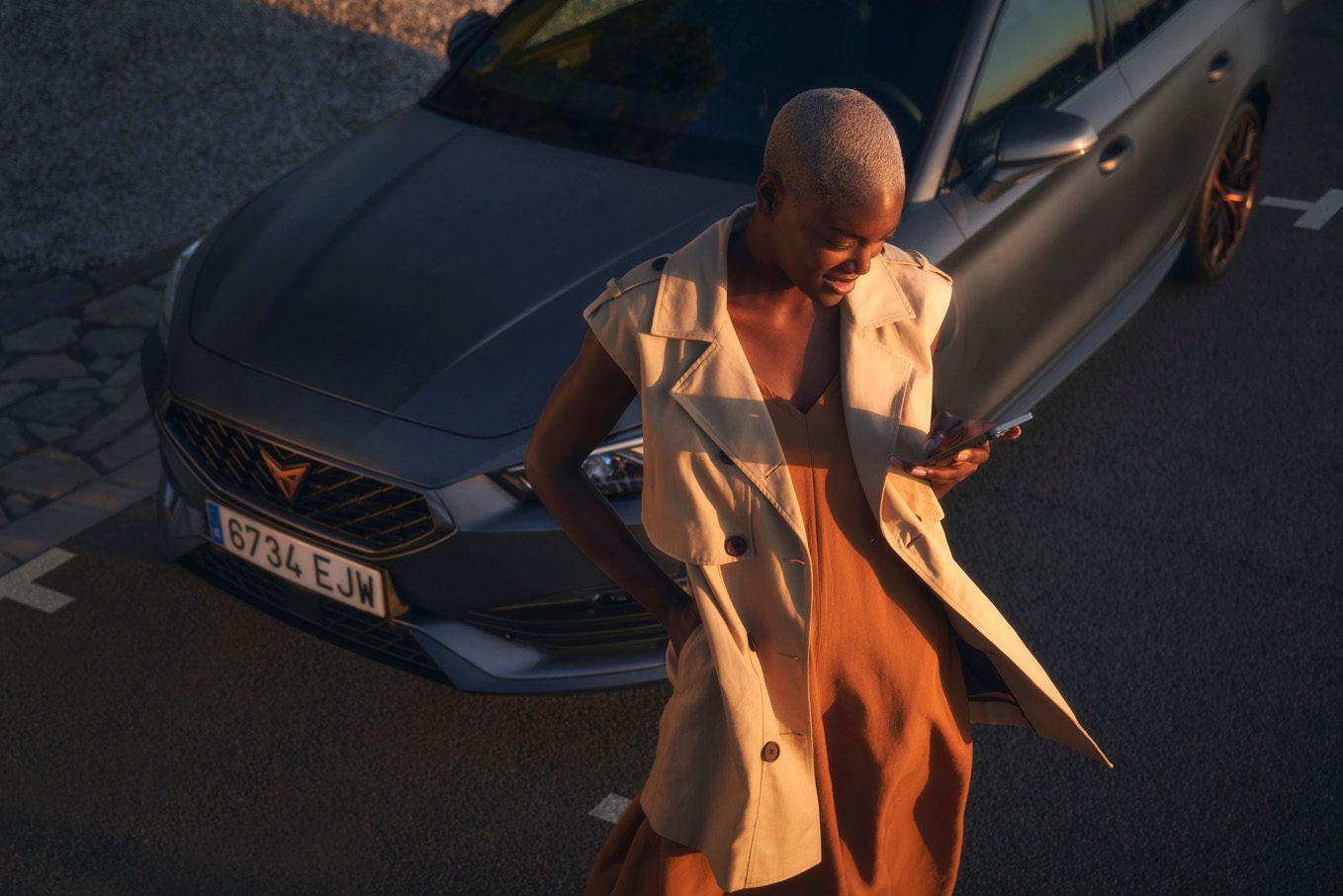 SUV stojí na parkovacom mieste na ceste. Od vozidla odchádza žena, ktorá drží mobilný telefón a pozerá sa na neho. Oblečené má oranžové šaty a béžovú vestu.