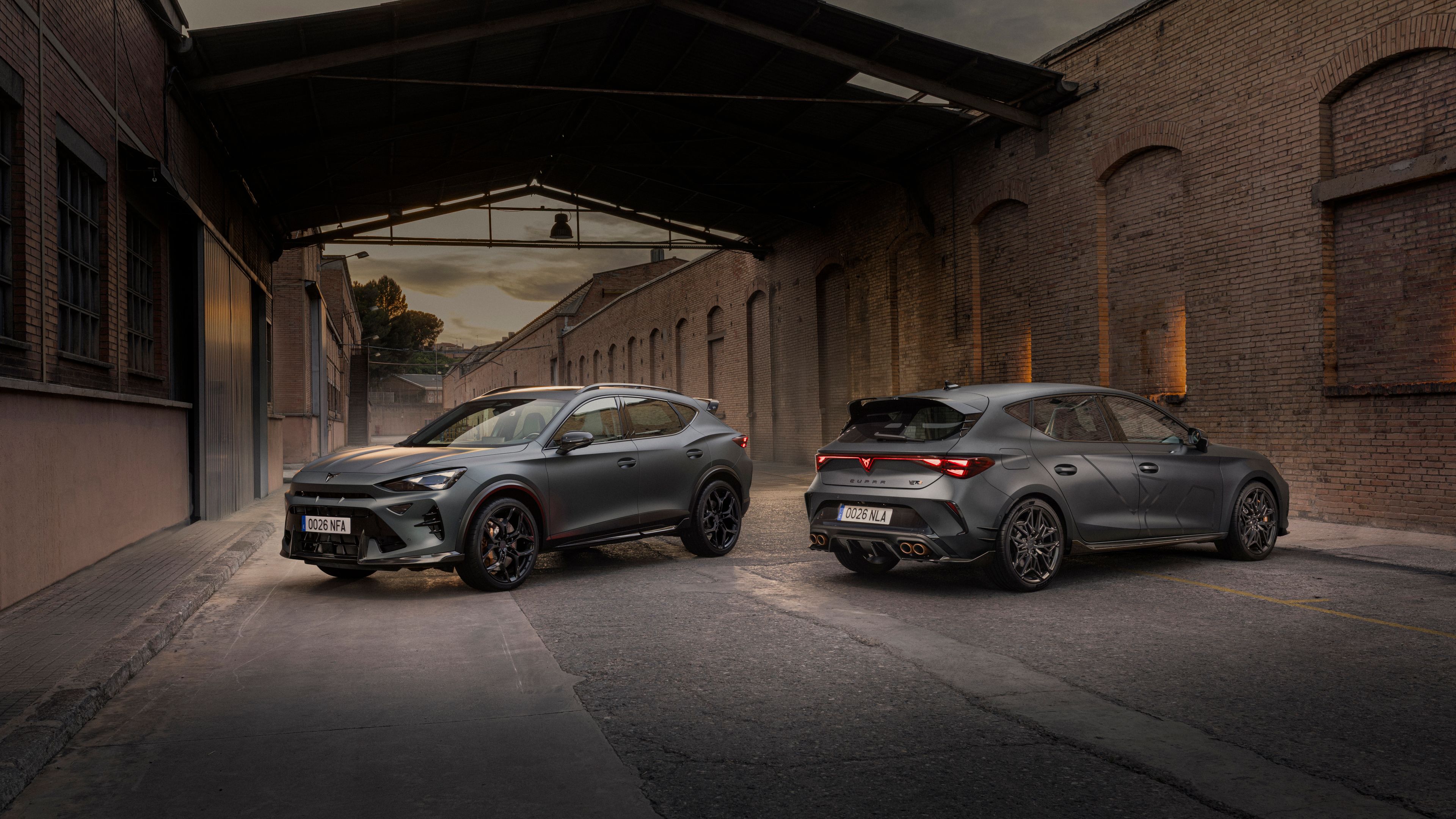 Modely CUPRA zaparkované na ceste medzi dvoma priemyselnými budovami za súmraku.