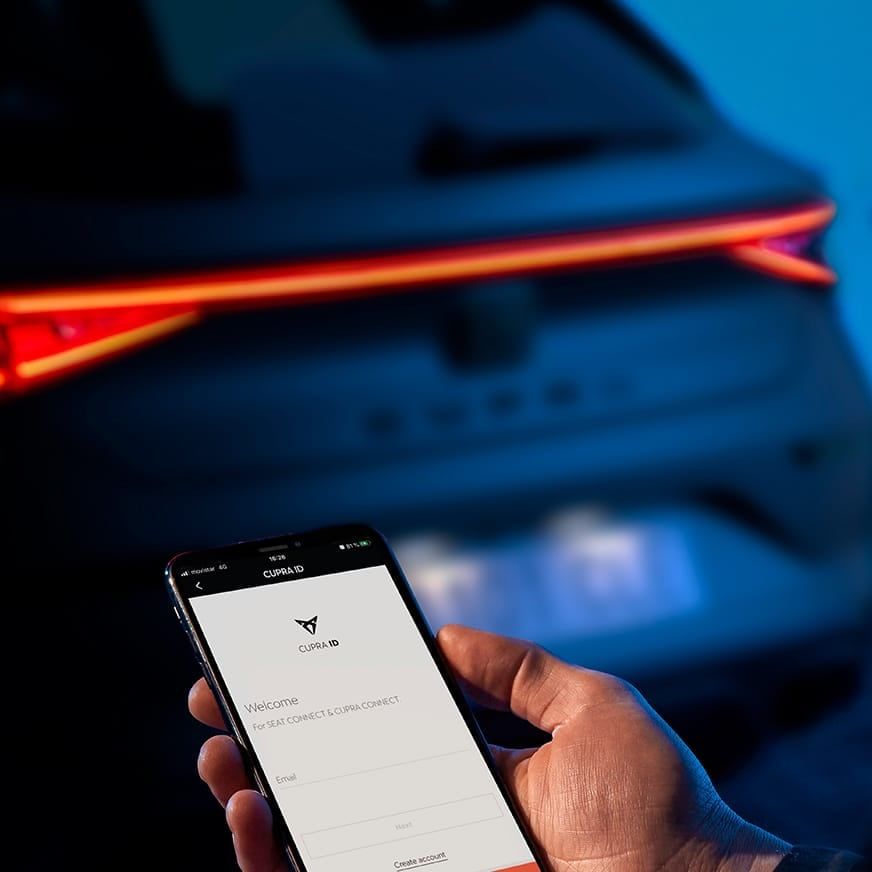 Ruka drží mobilný telefón s otvorenou aplikáciou CUPRA. Svetlo auta svieti jasne červenou farbou. Pozadie tvorí rozostrené vozidlo.