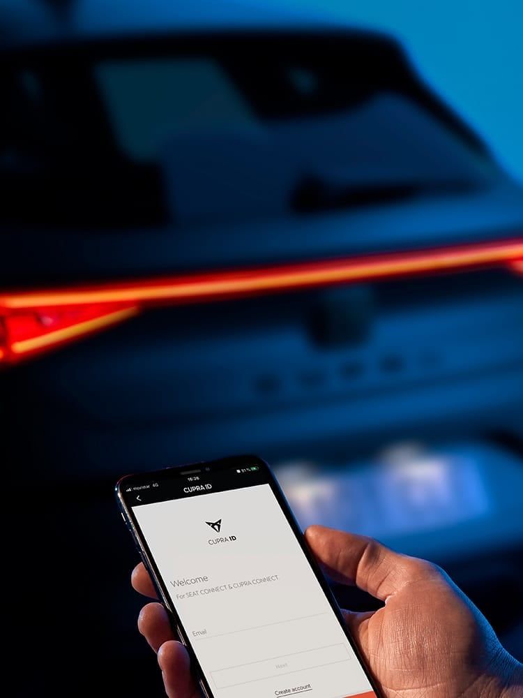 Ruka drží mobilný telefón s otvorenou aplikáciou CUPRA. Svetlo auta svieti jasne červenou farbou. Pozadie tvorí rozostrené vozidlo.