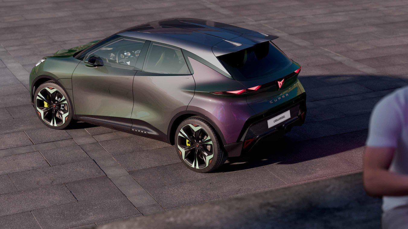 CUPRA UrbanRebel s moderným dizajnom a športovými líniami zaparkované na dláždenom povrchu. Farba auta má gradientný efekt s odtieňmi fialovej a zelenej. V popredí je muž v bielom tričku.