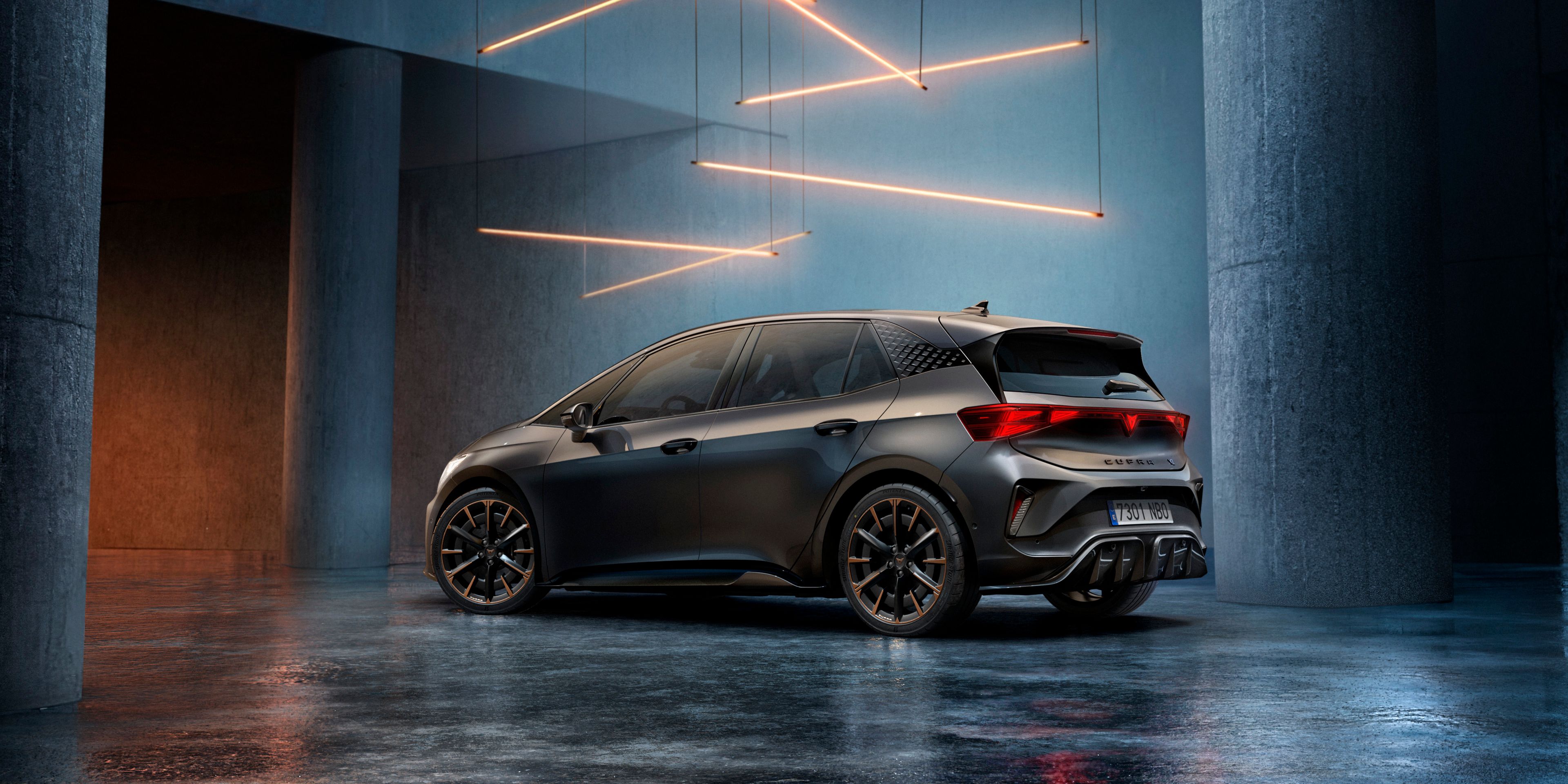 CUPRA Born stojí v štýlovo osvetlenom priemyselnom priestore s betónovými stenami a stĺpmi. Zo stropu visí niekoľko lineárnych svetelných zdrojov, ktoré vytvárajú elegantnú atmosféru. Vozidlo je vidieť zo zadnej strany a odráža svetlo na lesklej podlahe.
