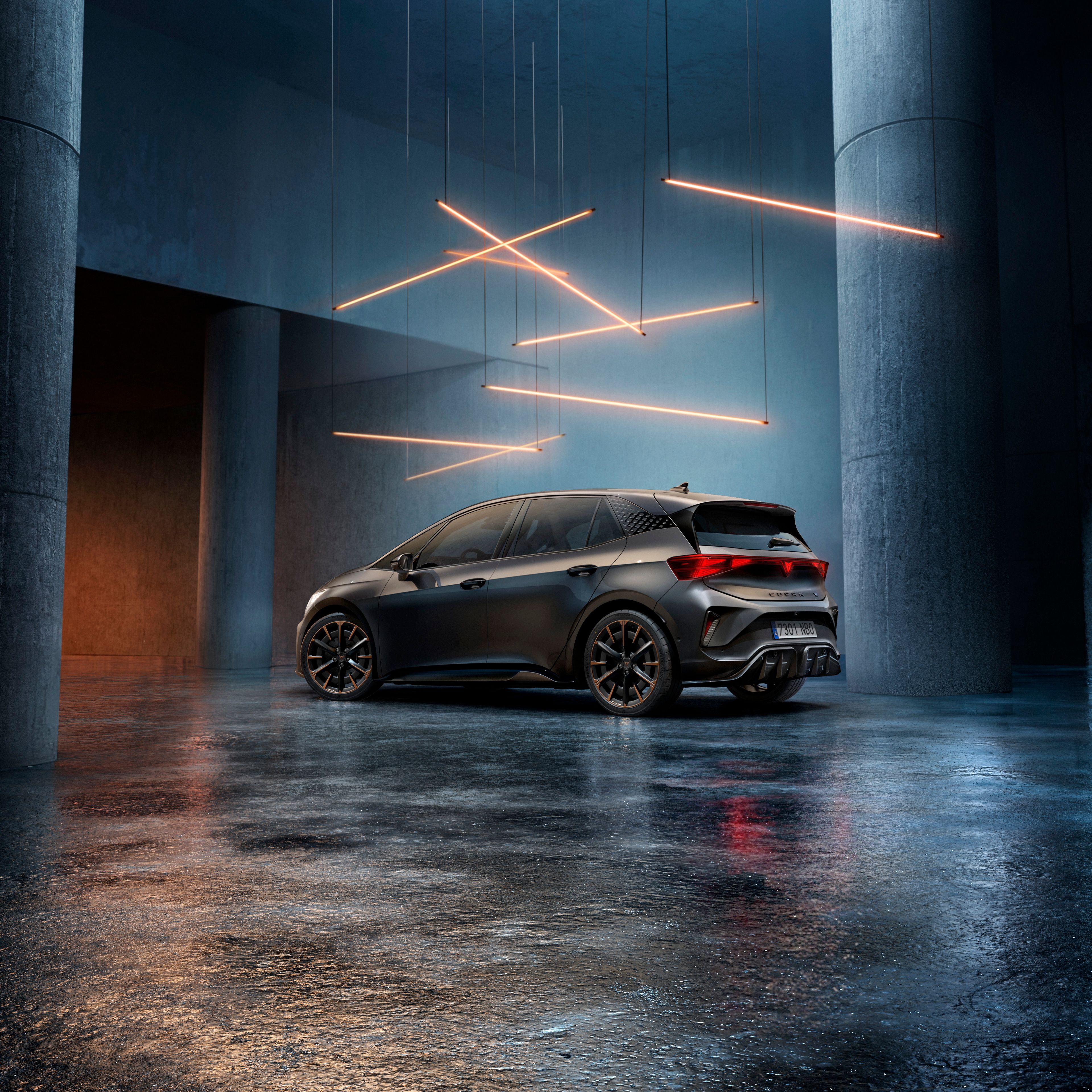 CUPRA Born stojí v štýlovo osvetlenom priemyselnom priestore s betónovými stenami a stĺpmi. Zo stropu visí niekoľko lineárnych svetelných zdrojov, ktoré vytvárajú elegantnú atmosféru. Vozidlo je vidieť zo zadnej strany a odráža svetlo na lesklej podlahe.