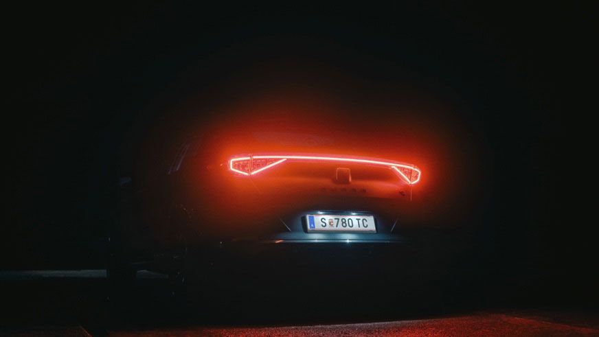 Zadná časť tmavého auta s rozsvietenými červenými zadnými LED svetlami, odrážajúca sa v mokrom povrchu cesty. Pozadie je tmavé a vytvára dramatickú atmosféru.