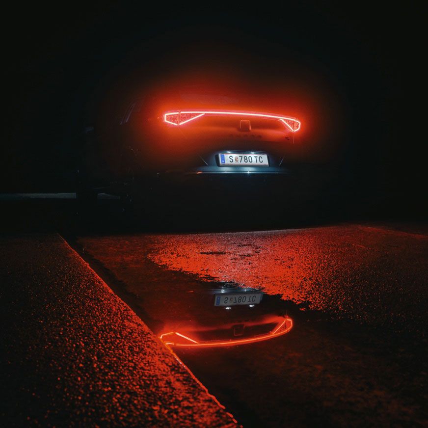 Zadná časť tmavého auta s rozsvietenými červenými zadnými LED svetlami, odrážajúca sa v mokrom povrchu cesty. Pozadie je tmavé a vytvára dramatickú atmosféru.